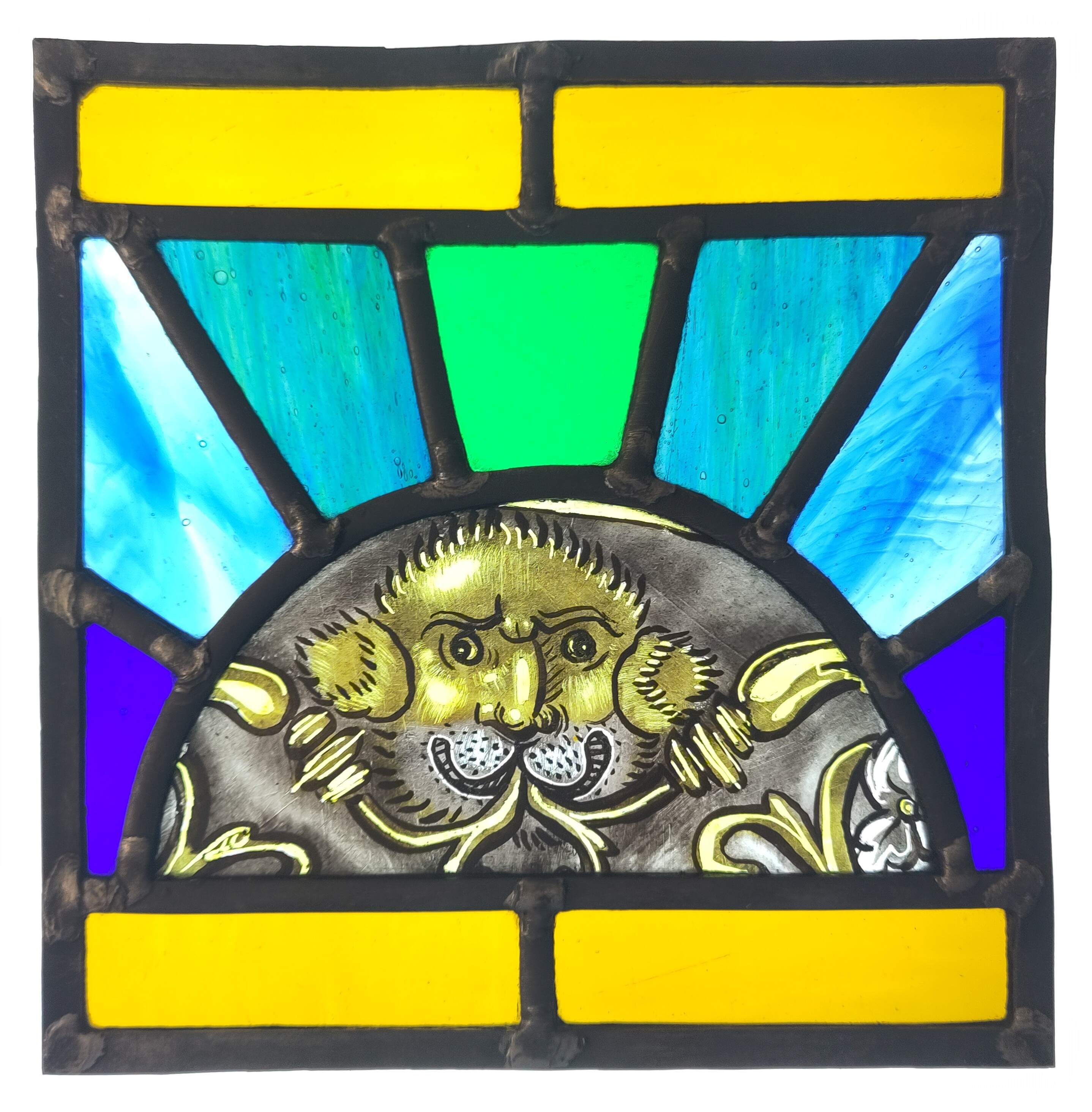 An image of a stained glass panel. The centre piece of the panel is a semi-circular piece with a painted lion-like grotesque figure with foliage decoration, all coloured yellow. This painted piece is surrounded by green and blue glass radiating out from the cntre, like a stylised sun. The panel is illuminated from the back.